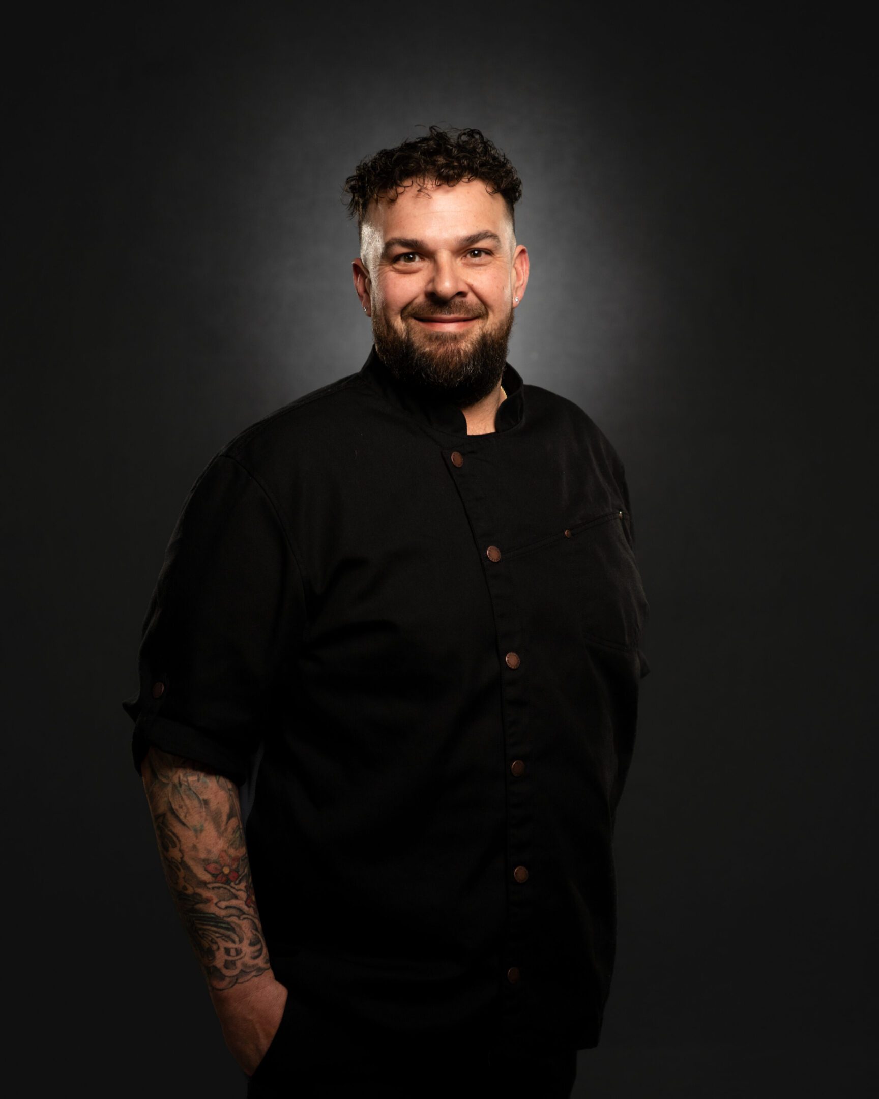 Smiling man with beard and tattoos wearing a black shirt against a dark background.