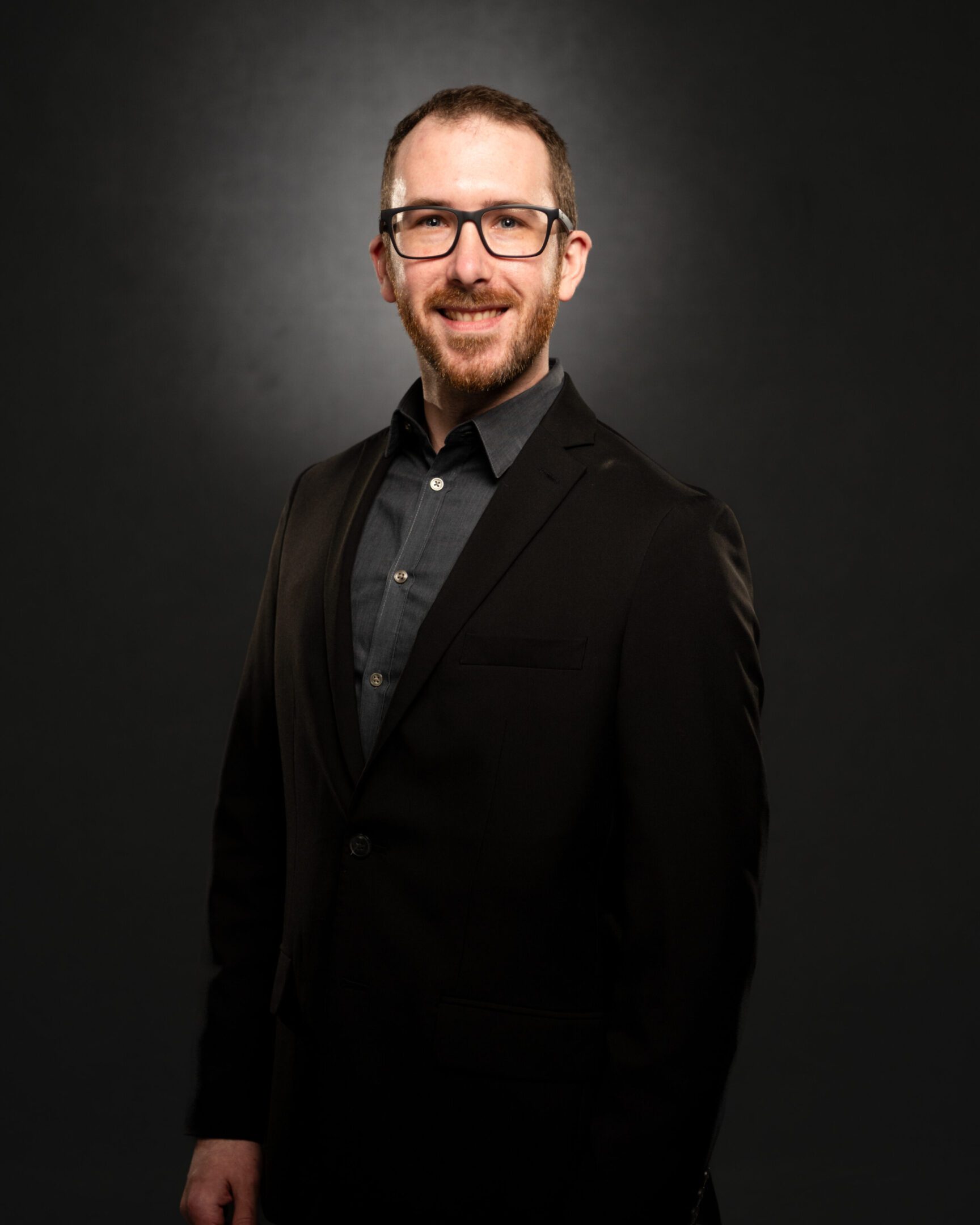 Smiling man in glasses wearing a dark suit against a black background.