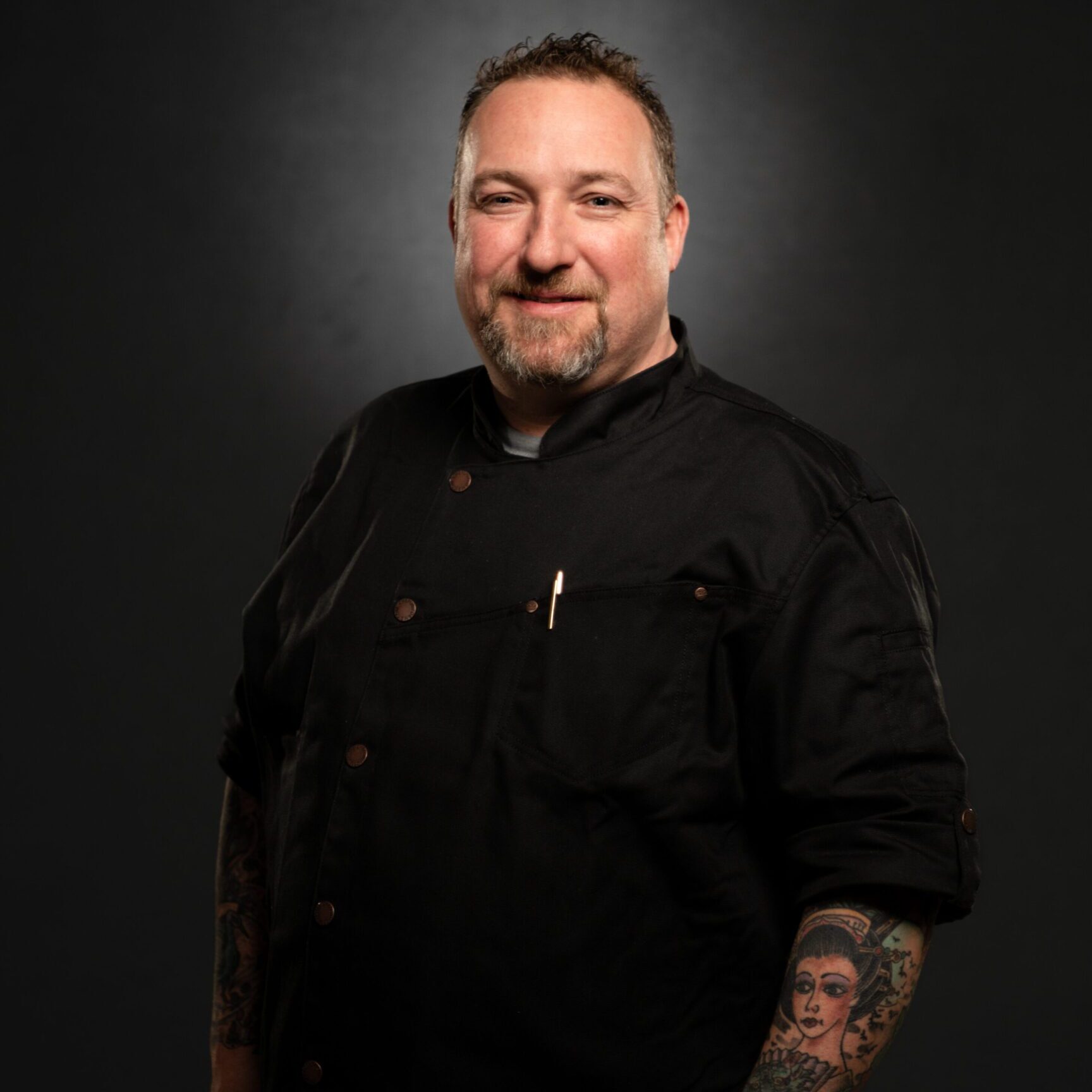 Chef in a black uniform with tattooed arms posing against a dark background.